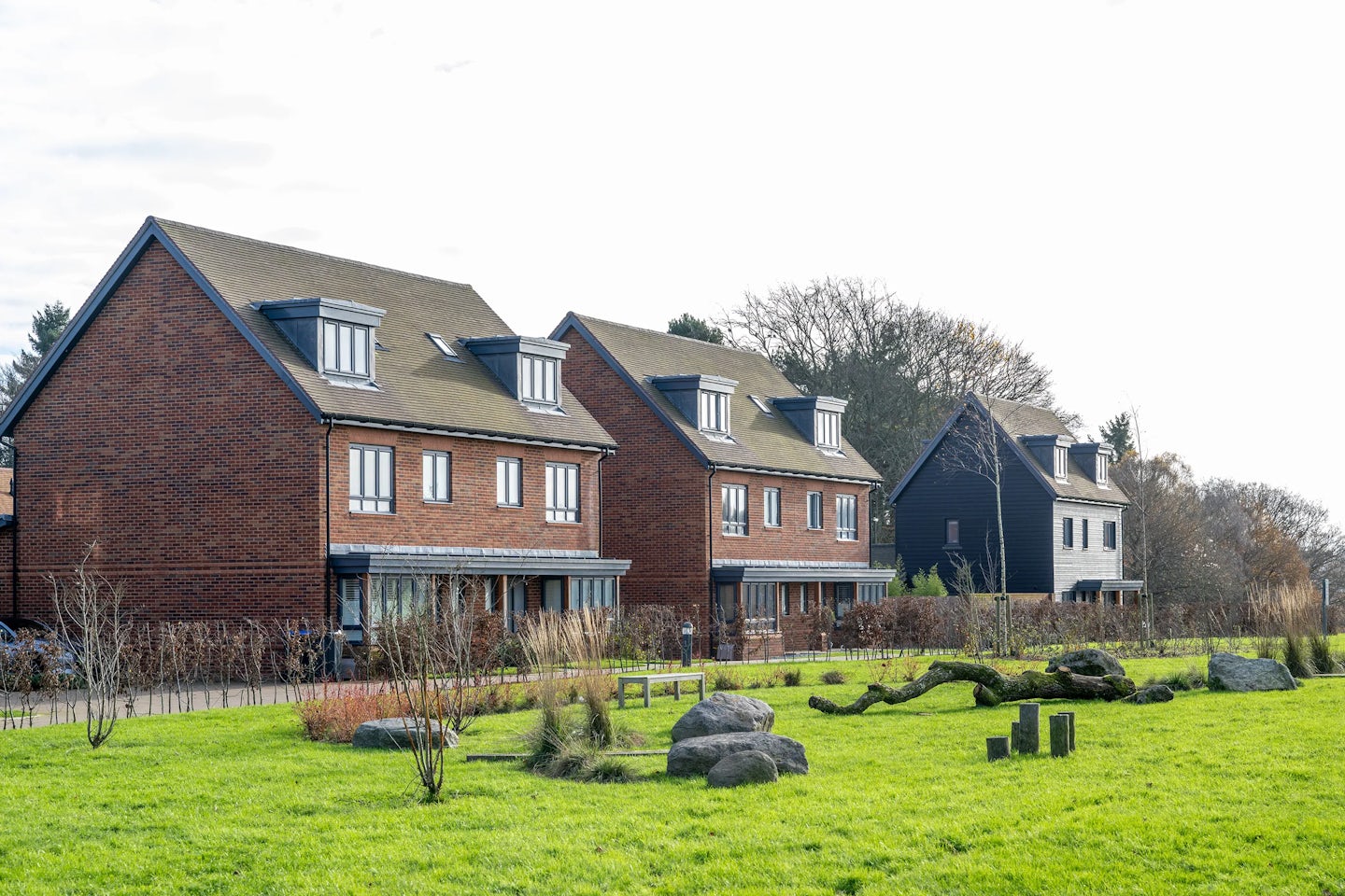 A row of semi detached brick houses with bay windows and dormer windows in the roof line the edge of a large green open space. Rocks and decorative items feature in the open area.