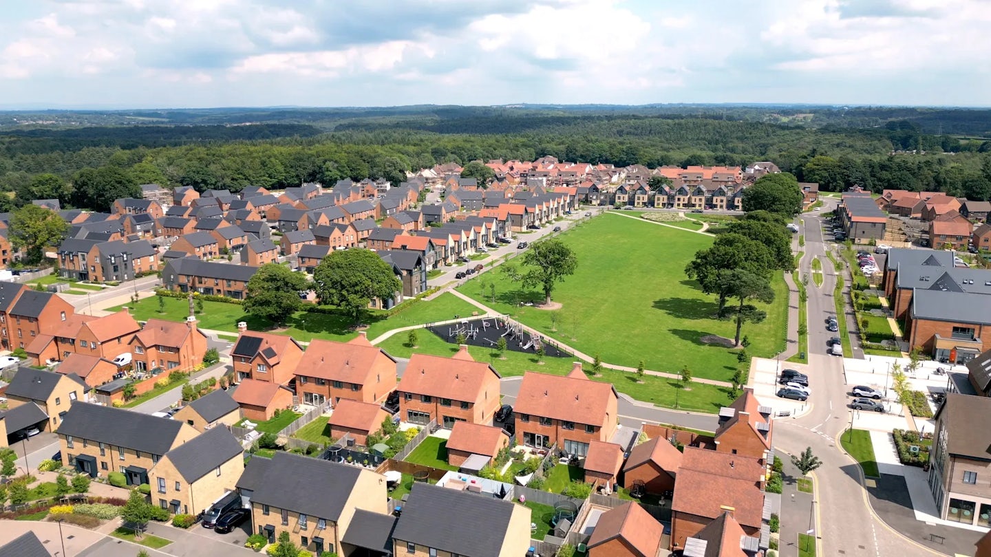 An aerial view of Woodgate at Pease Pottage, show the large village green lines by nordic style villas and traditional sussex style brick houses. The community hub cafe and shop along with St Catherines hospice are in the bottom right of the green.