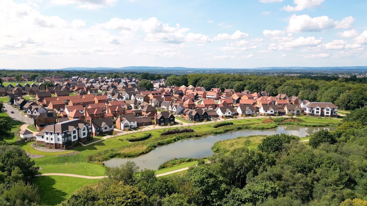 An aerial view of Woodgate at Pease Pottage, showing the large lake, running path, forest, and collection of Sussex style houses.