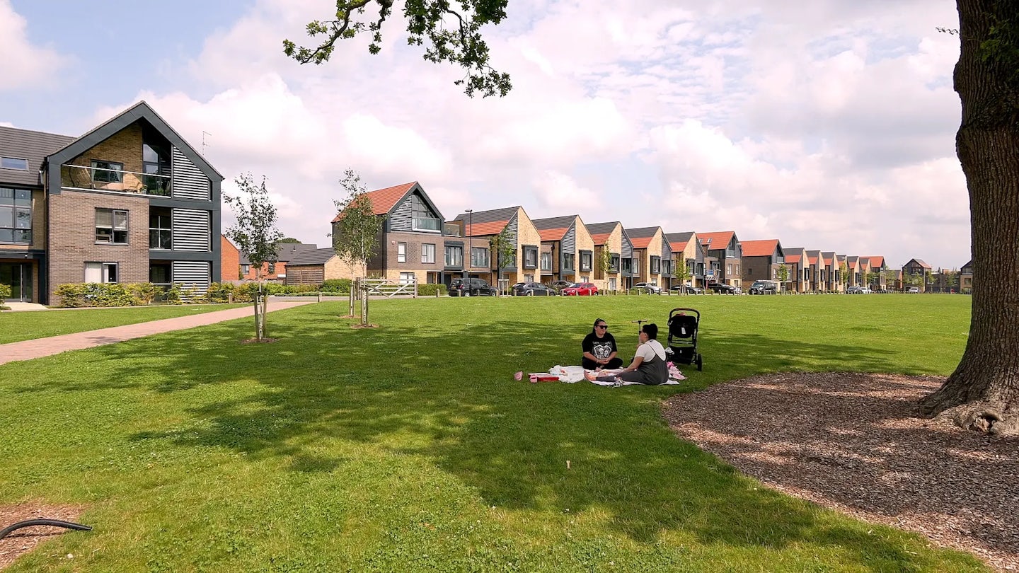 People site in the ahde of a mature tree in the large village green of Woodgate at Pease Pottage. Nordic style villas can be seen in the background lining the edge of the green.