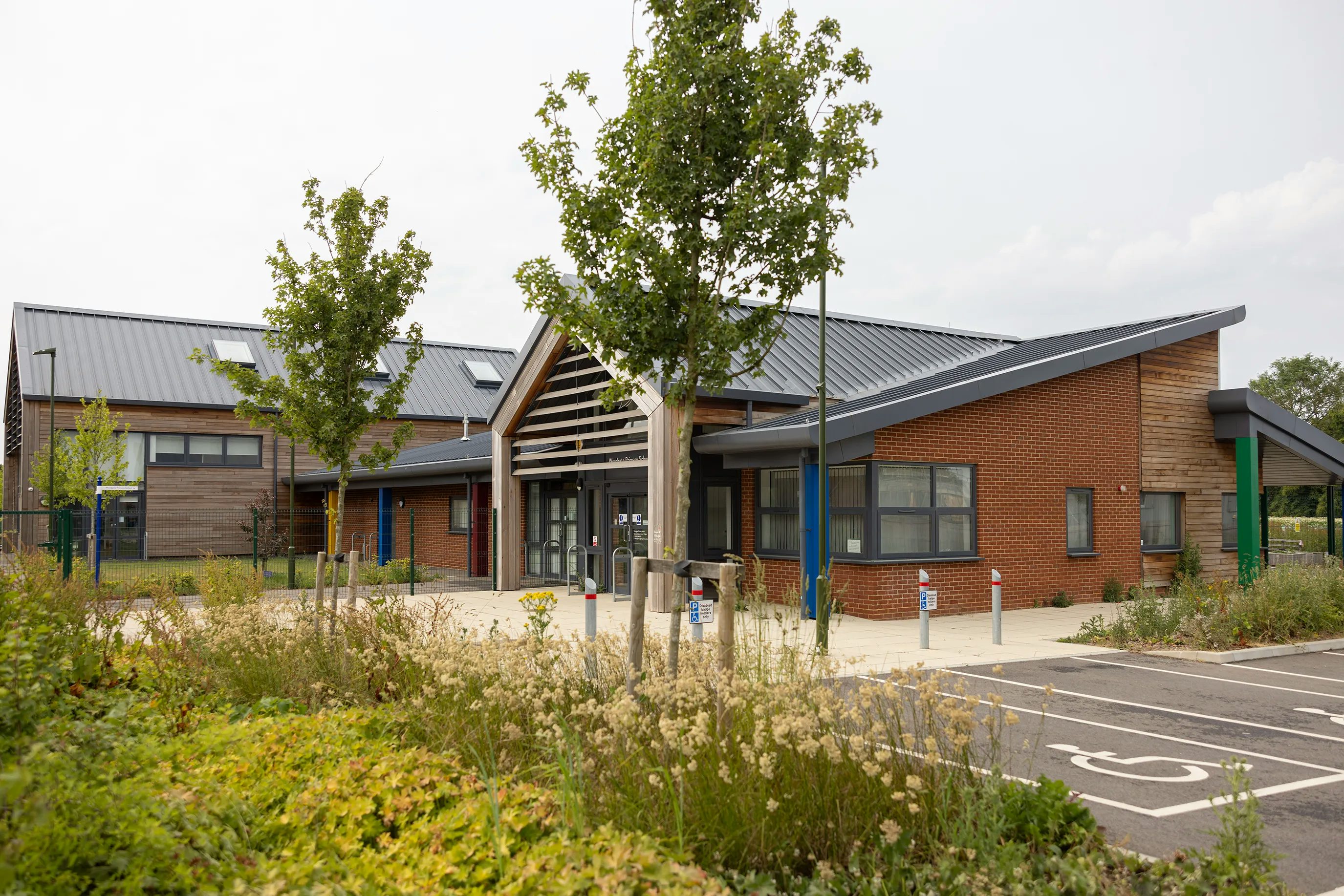 A side view of the entrance to Woodgate Primary School. The side is brick, the roof is grey, and the entrance has a wooden frame in nordic design.