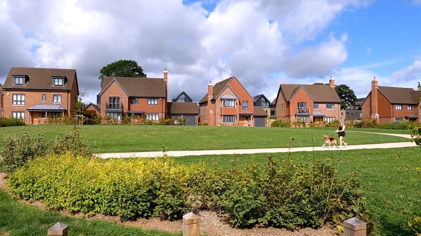 A woman walks a dog along a gravel path in a large green area. A row of new build brick homes site behind the green space.