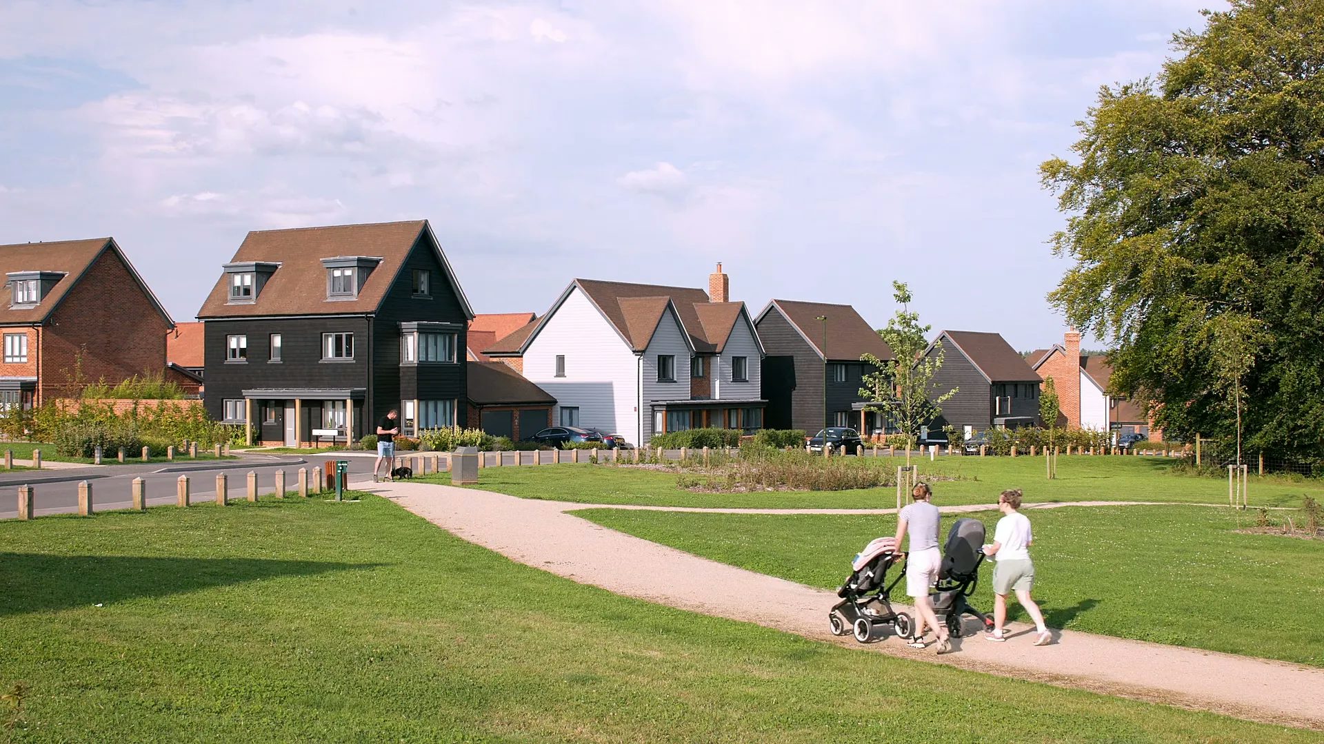 A couple with push chairs walk along a gravel path with a row of new build homes in front of them. A man walks his dog in the opposite direction. The houses are clad in black and white.