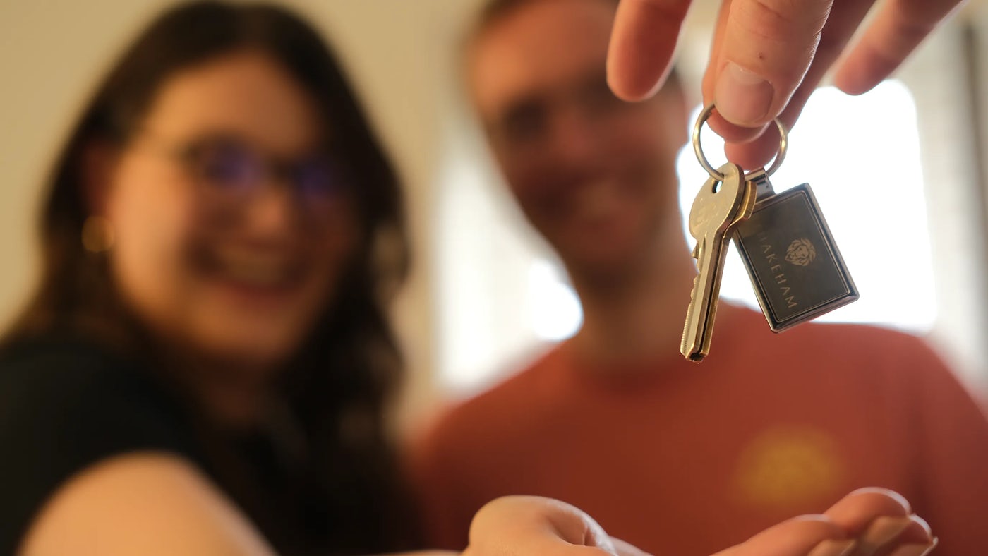 A couple are out of focus in the background but are smiling, a woman with pinkish nail polish holds out a hand to collect some keys which are being handed from above.