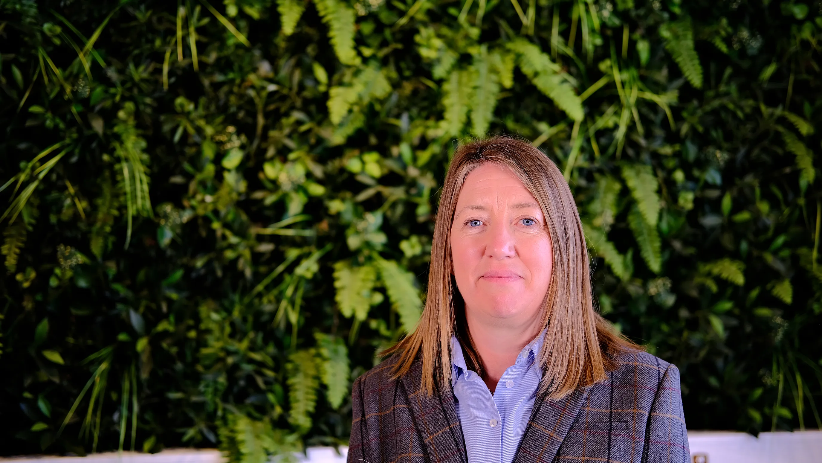 Mintha wears a blue shirt and tweed jacket. She has light brown hair to her shoulders. She sits in front of green foliage.