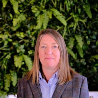 Mintha wears a blue shirt and tweed jacket. She has light brown hair to her shoulders. She sits in front of green foliage.