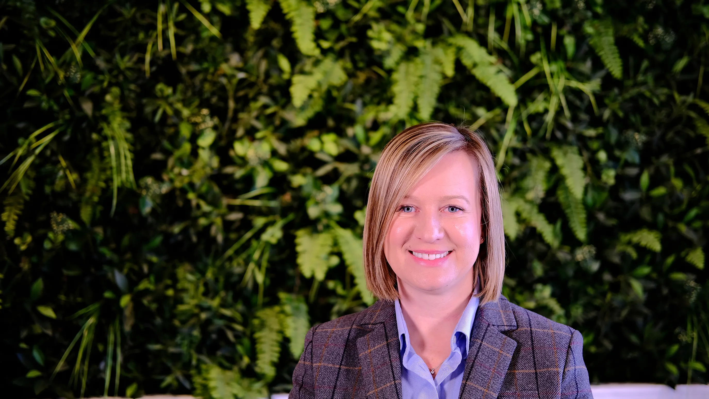 Sarah wears a blue shirt and tweed jacket. She has blonde hair cut just above her shoulders. She sits in front of green foliage.