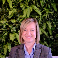 Sarah wears a blue shirt and tweed jacket. She has blonde hair cut just above her shoulders. She sits in front of green foliage.