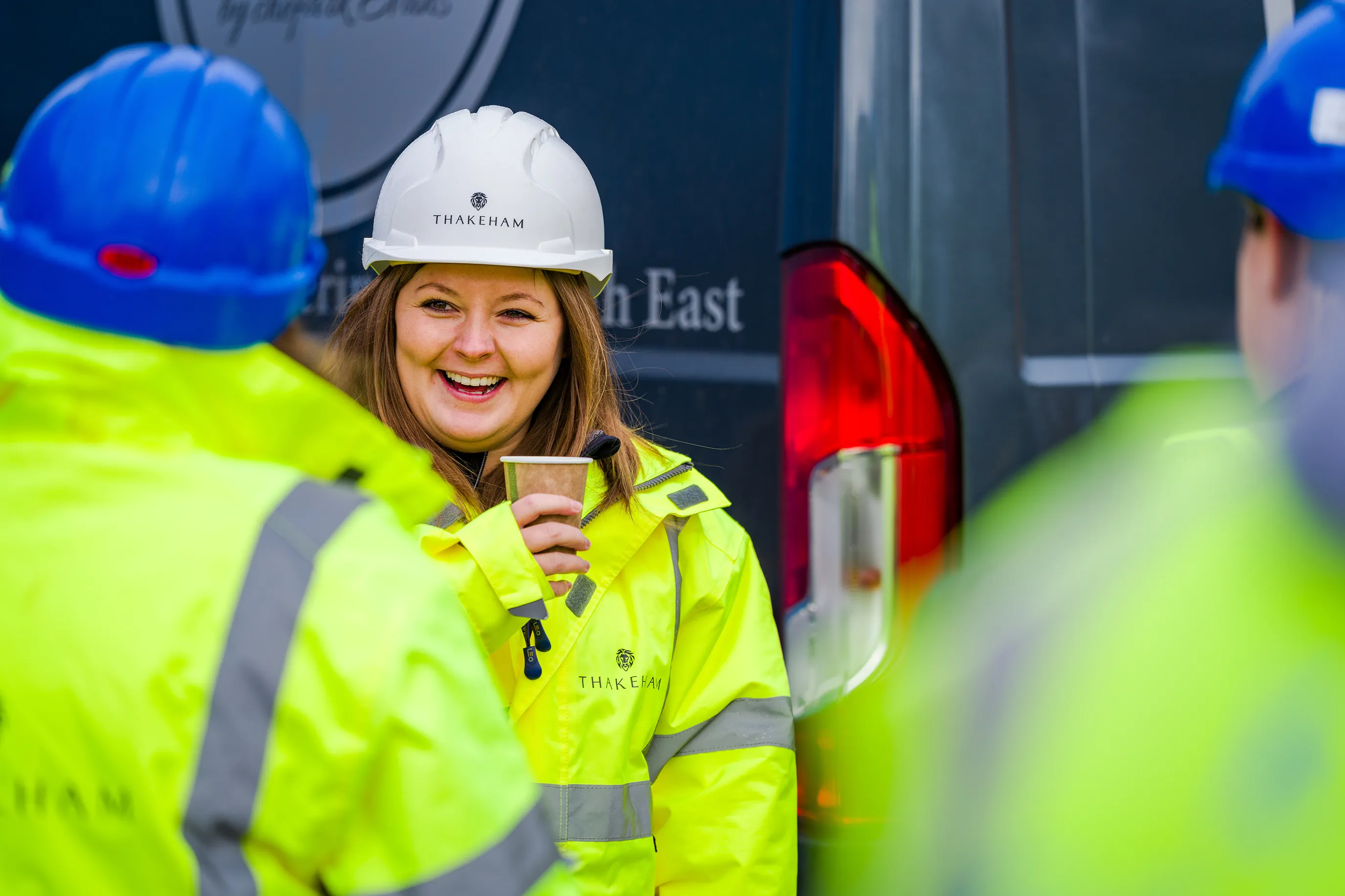 A group of construction workers enjoy a tea break. All are wearing high vis jackets and hard hats. One faces the camera holding a coffee