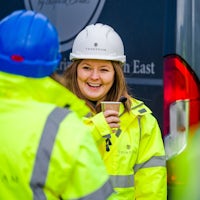A group of construction workers enjoy a tea break. All are wearing high vis jackets and hard hats. One faces the camera holding a coffee