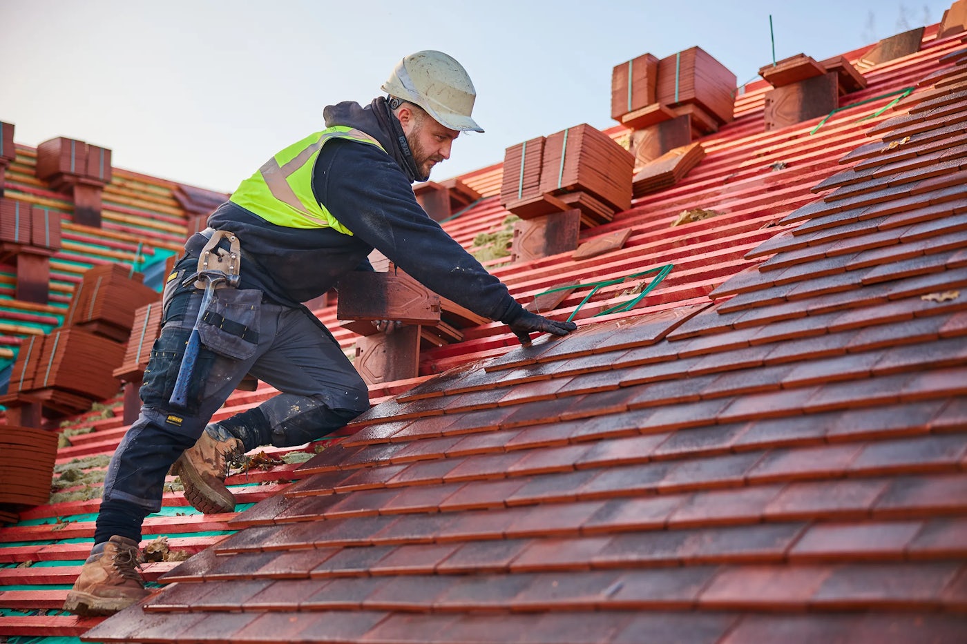 A construction worker in a high vis jacket and hard hat fixes tiles to a roof.