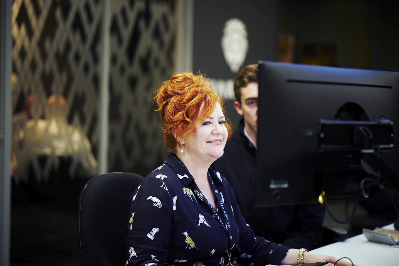 An office worker with red hair sits at a desk looking at a screen.