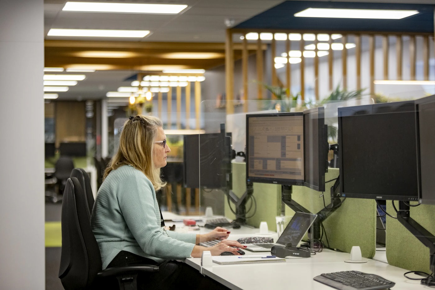 Office worker sits at a desk looking at a dual screen. Wider office layout can be seen behind them.