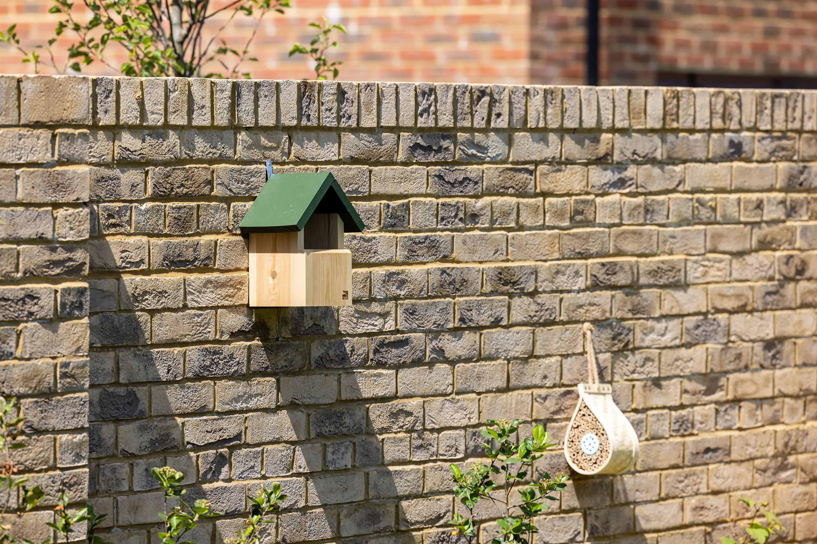 A bird box is fixed to a brick wall in a residential garden. Its made of wood with a green roof.