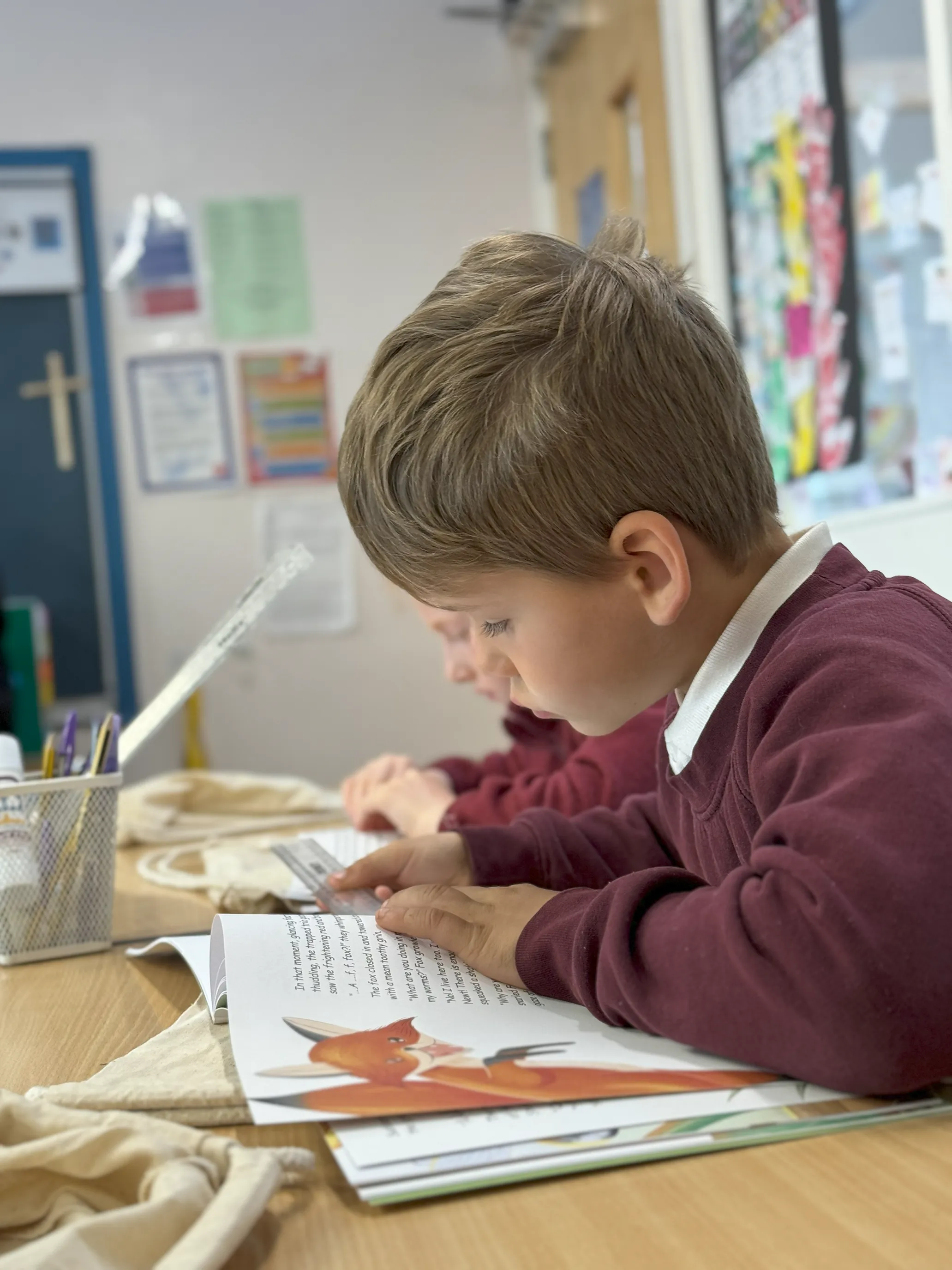 a young child in burgundy school uniform sitting in a classroom reads a book with a fox illustration..