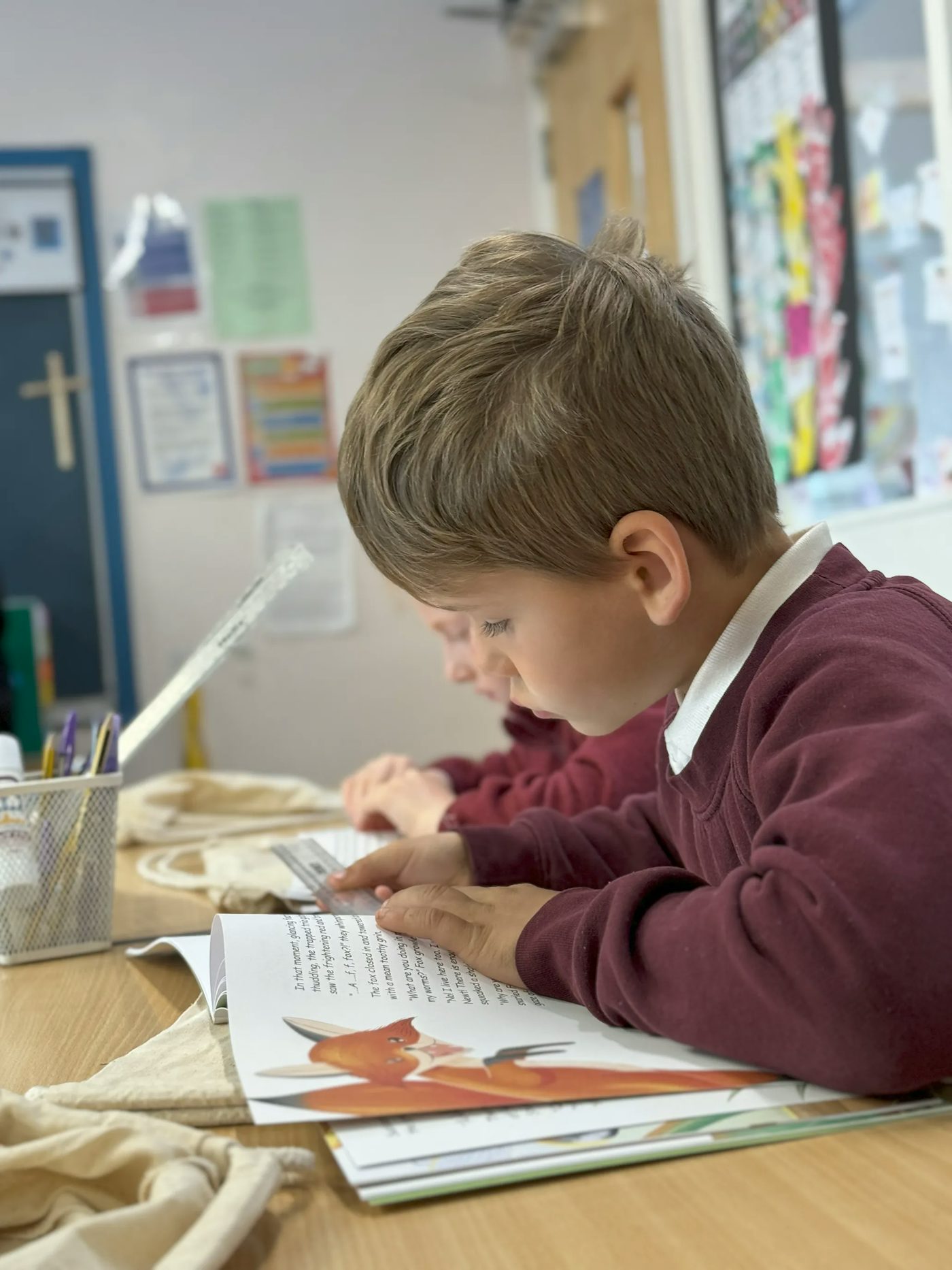 a young child in burgundy school uniform sitting in a classroom reads a book with a fox illustration..