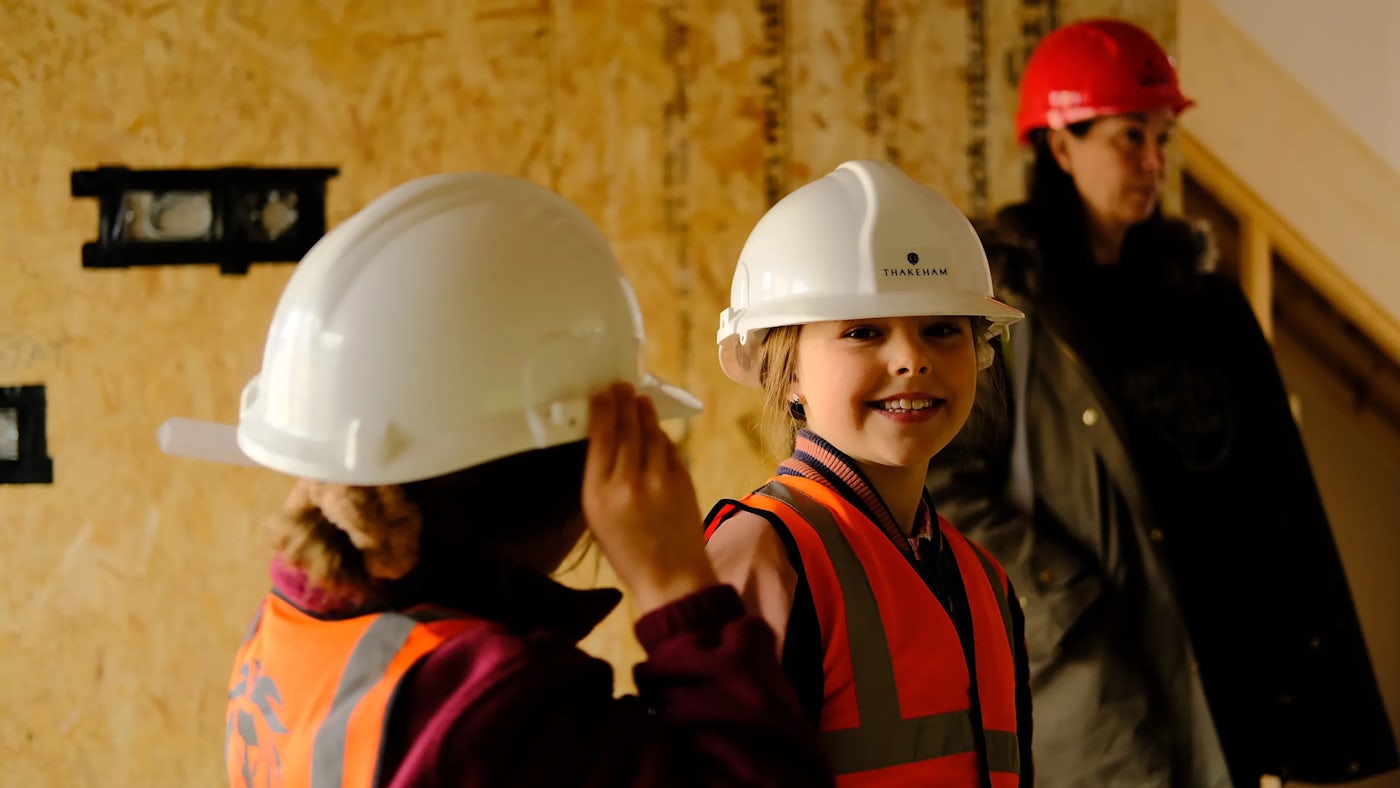a child wears a Thakeham branded hard hat and an orange high vis jacket.