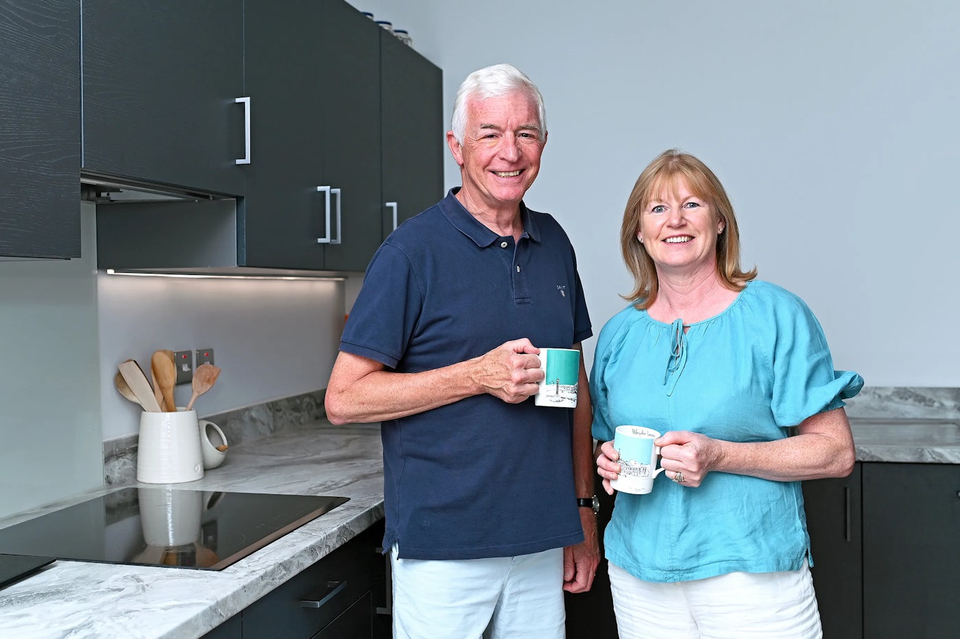 Jon and Sarah stand in their new kitchen at Manorwood in Surrey. Black cabinets and a light grey countertop are visible, included integrated appliances.
