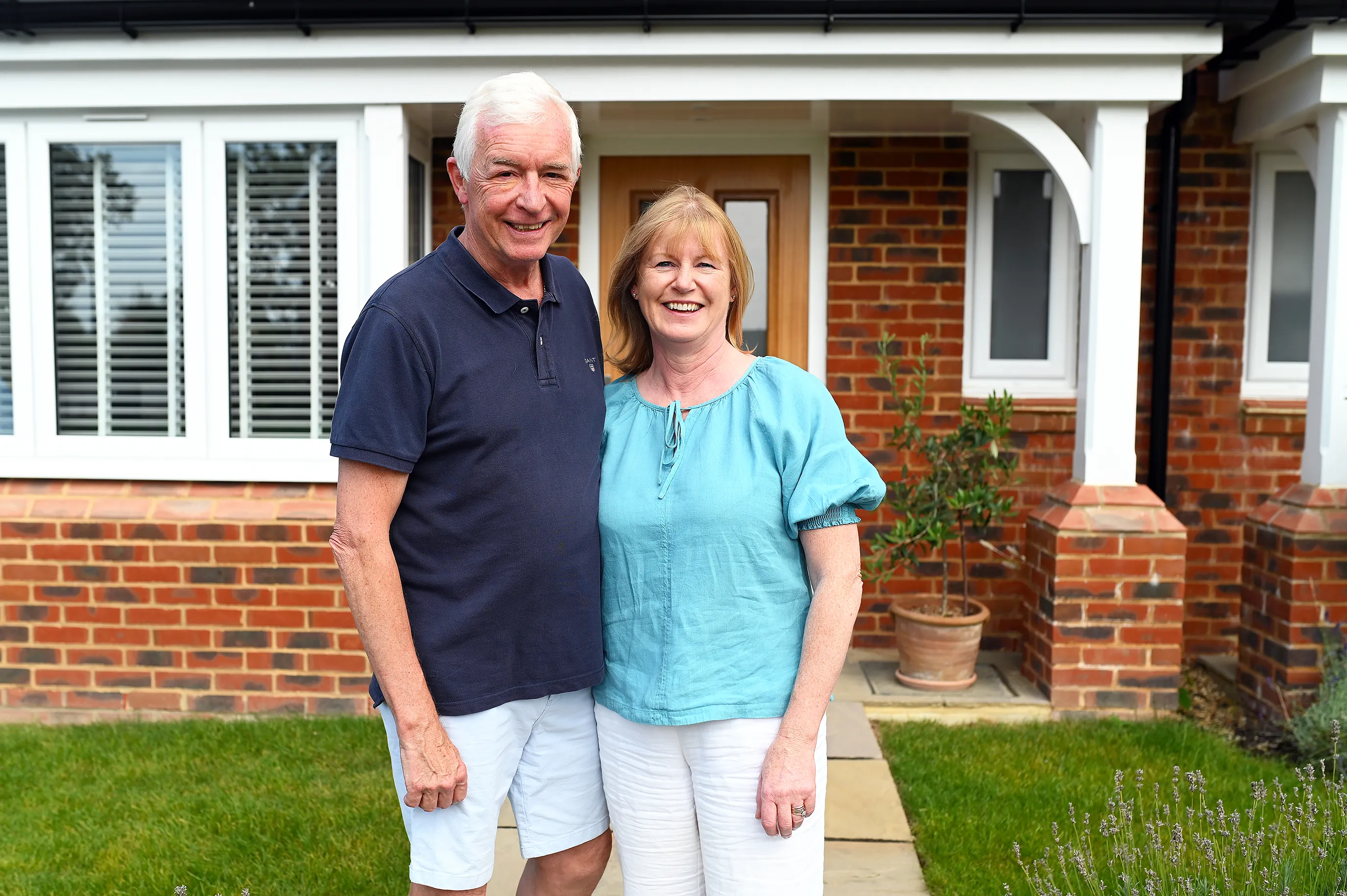 Jon and Sarah stand outside their new build home at Manorwood in Surrey. The ground floor of a brick house can be seen behind them.