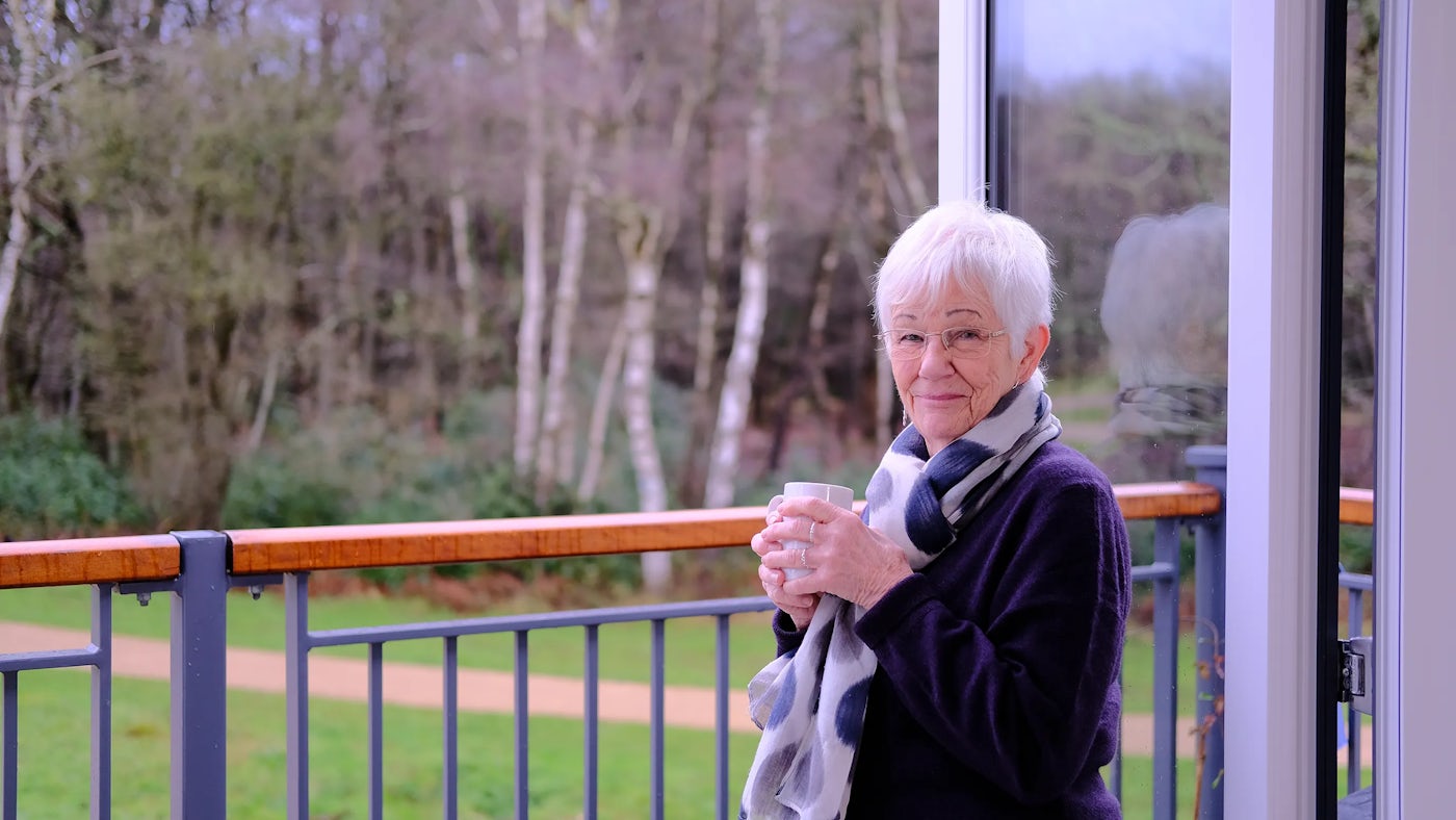 Jenny enjoy a cup of tea on the balcony of her new apartment at Woodgate. She stands facing the camera, forest and greenspace is visible behind her.