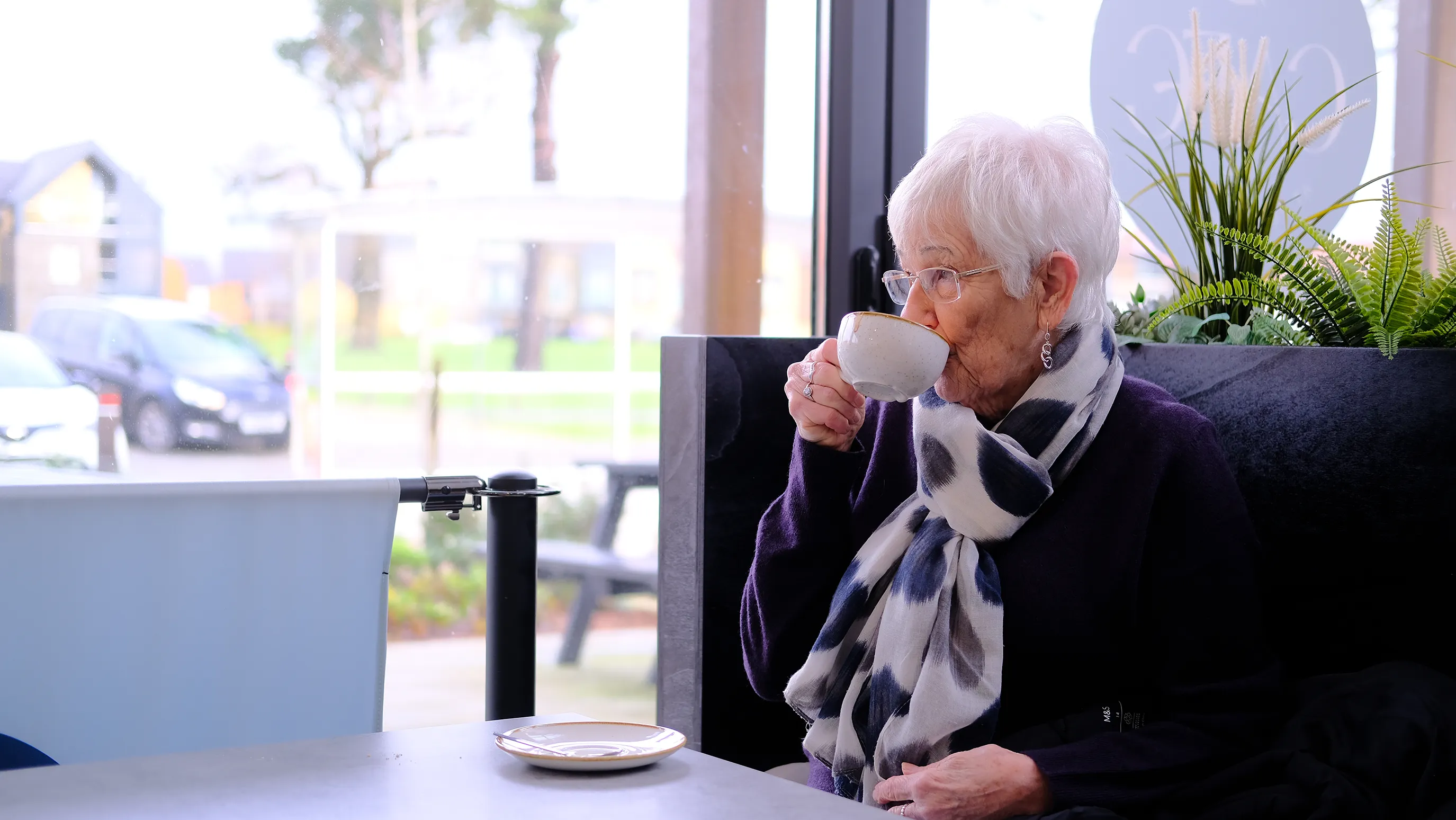 Jenny enjoys a coffee in the Woodgate cafe at Pease Pottage. She wears a white sild scarf with dark blue spots.