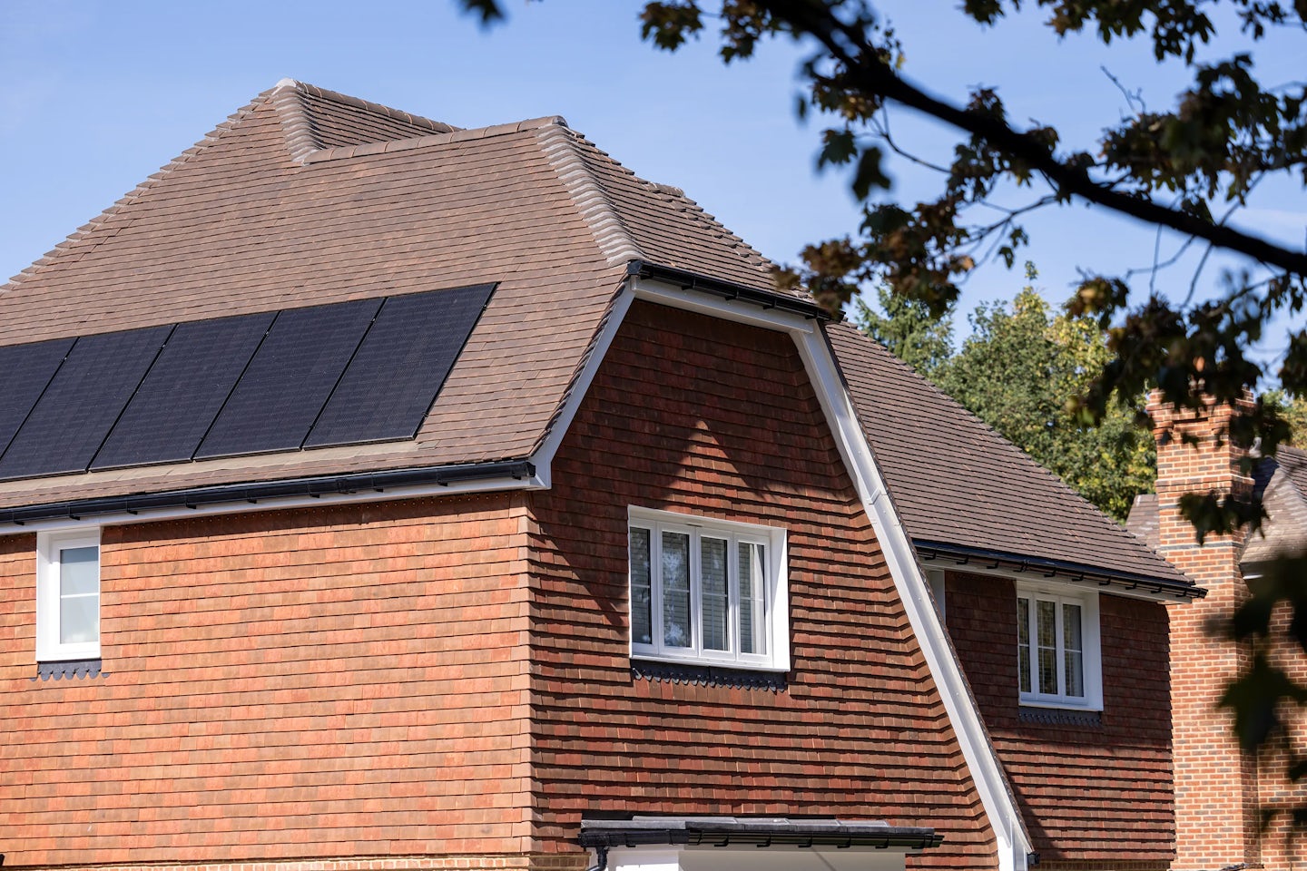 A pitched-roof house with brown shingles, featuring several solar panels installed on the roof. The house has brick walls and white-framed windows. The solar panels highlight the home's use of renewable energy.