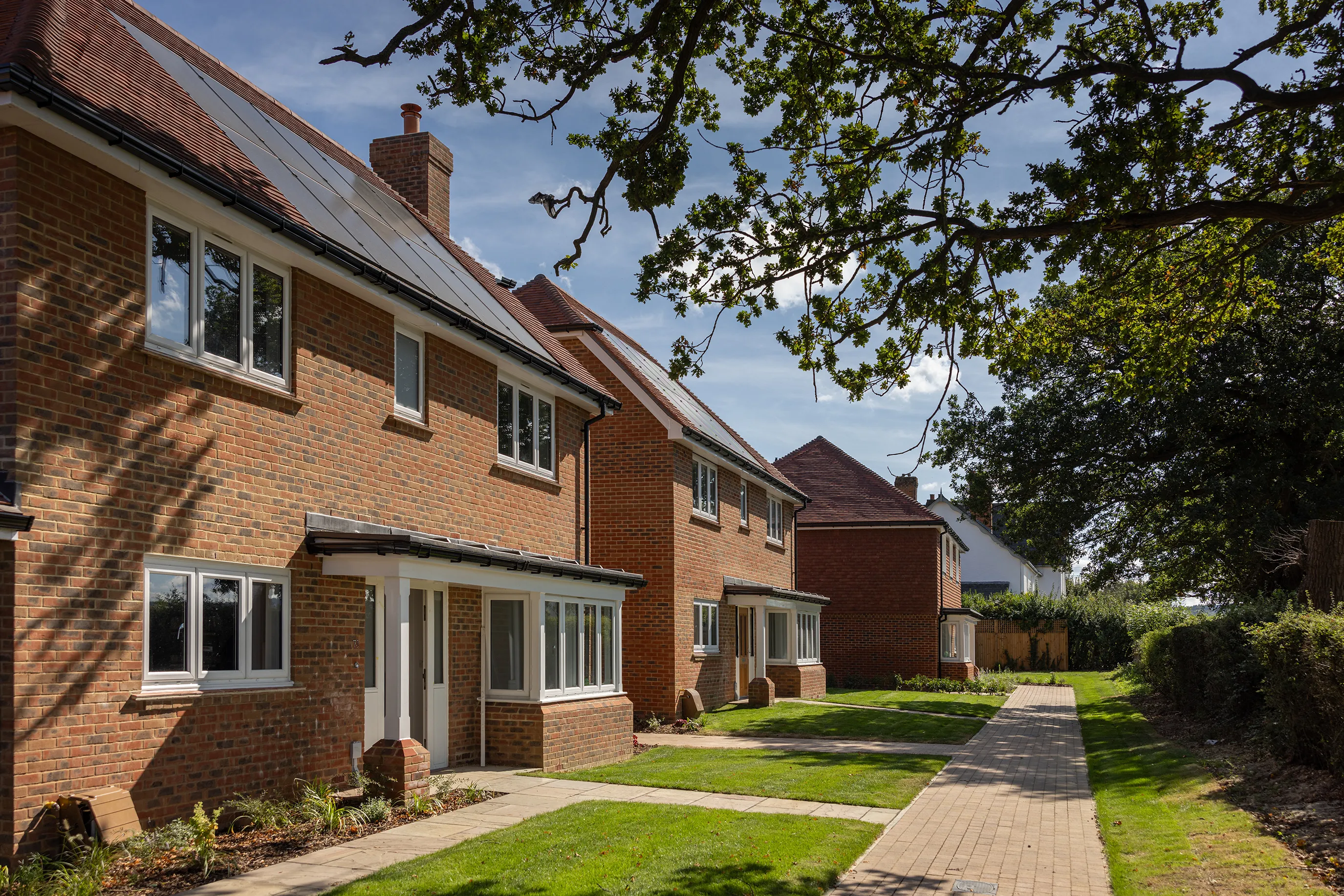 A row of modern, two-story brick houses with well-maintained lawns and a paved walkway in front. The houses have large windows, and some feature solar panels on their roofs. The scene is set on a sunny day with a clear blue sky, and trees provide some shade. The image highlights a suburban residential area with energy-efficient homes, emphasizing modern architectural design and sustainable living.