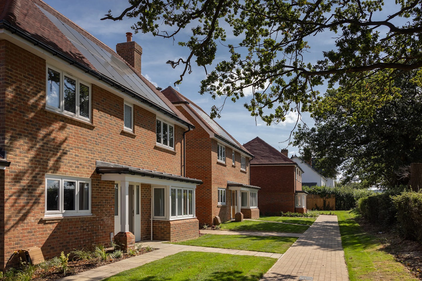 A row of modern, two-story brick houses with well-maintained lawns and a paved walkway in front. The houses have large windows, and some feature solar panels on their roofs. The scene is set on a sunny day with a clear blue sky, and trees provide some shade. The image highlights a suburban residential area with energy-efficient homes, emphasizing modern architectural design and sustainable living.