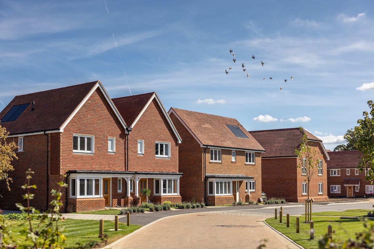 A suburban neighborhood featuring modern brick houses with pitched roofs, white-framed windows, and well-maintained front gardens. The houses include skylights, and the sky is mostly clear with a few scattered clouds. A flock of birds is flying in the distance, adding a dynamic element to the scene. The composition highlights a well-planned residential area with a neat, orderly aesthetic.