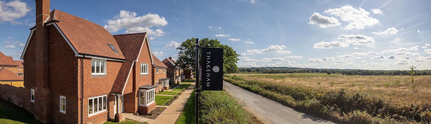 A row of modern brick houses with red roofs lines the left side of a paved walkway. A signpost reading "THAKEHAM" with a circular logo stands prominently. On the right, a road runs parallel to the houses, bordered by bushes, leading to an open field with grass and trees in the background. The sky is clear with scattered clouds, suggesting a sunny day. The setting captures a serene residential area blending nature with contemporary housing.
