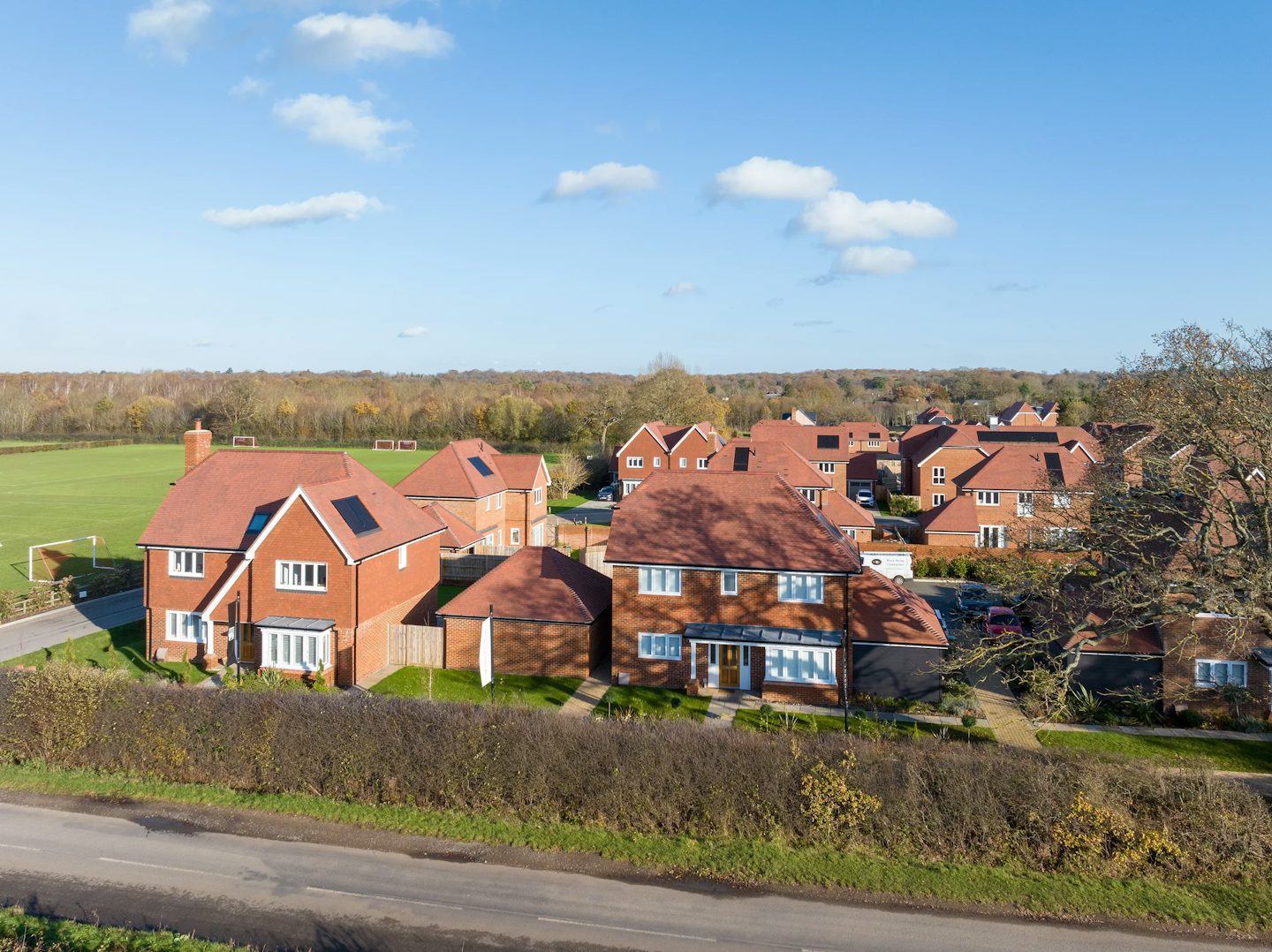 An aerial view of a suburban neighborhood featuring red-brick houses with red-tiled roofs, neatly arranged with well-maintained lawns and driveways. A road runs through the foreground, while a large green field extends into the background. The sky is mostly clear with a few scattered clouds.