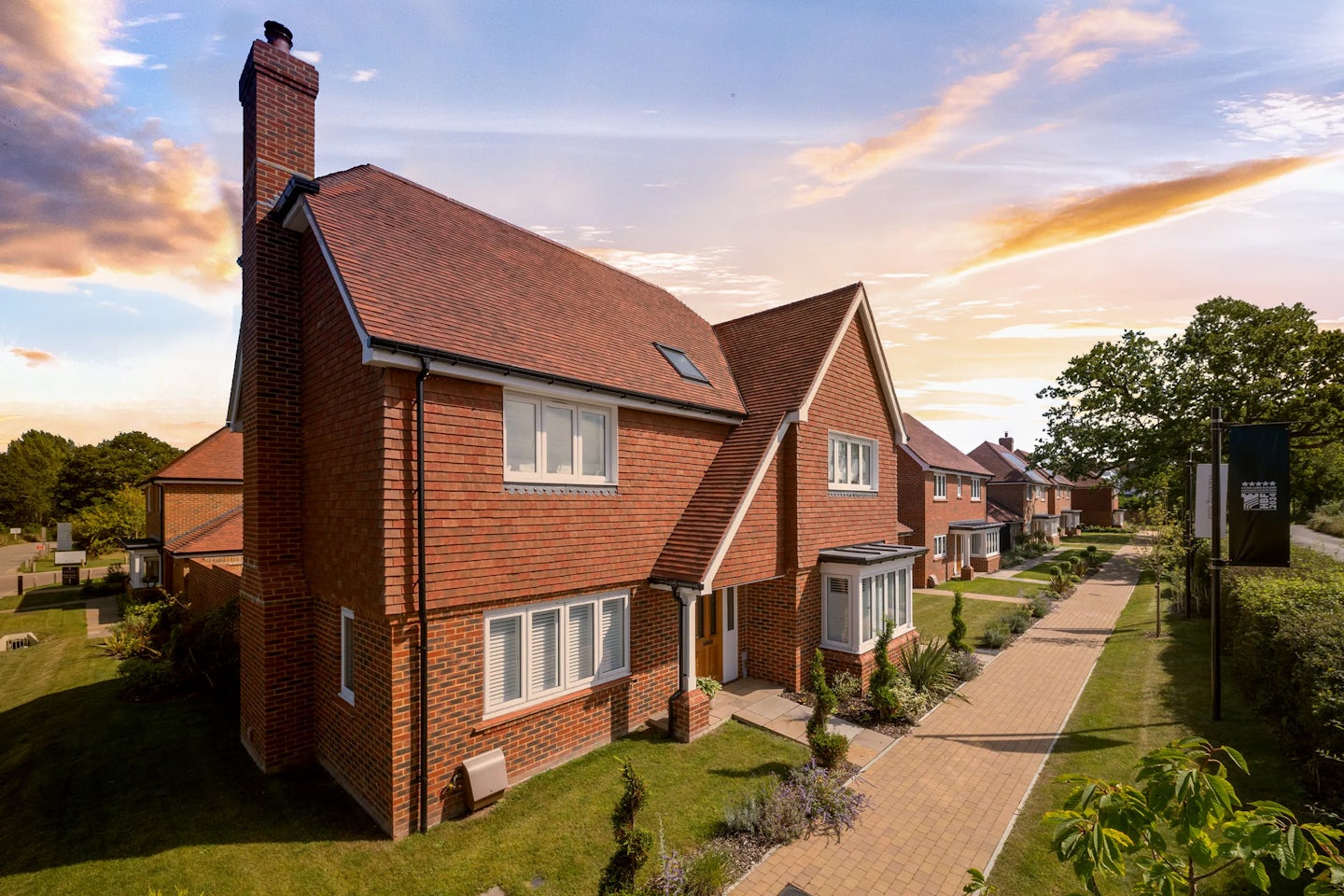 A row of modern detached houses in a suburban neighborhood. The houses have red brick exteriors, pitched roofs, and large windows. A well-maintained garden with shrubs and a paved walkway leads to the front door in the foreground. The sky is partly cloudy with a warm, golden hue, suggesting either sunrise or sunset.