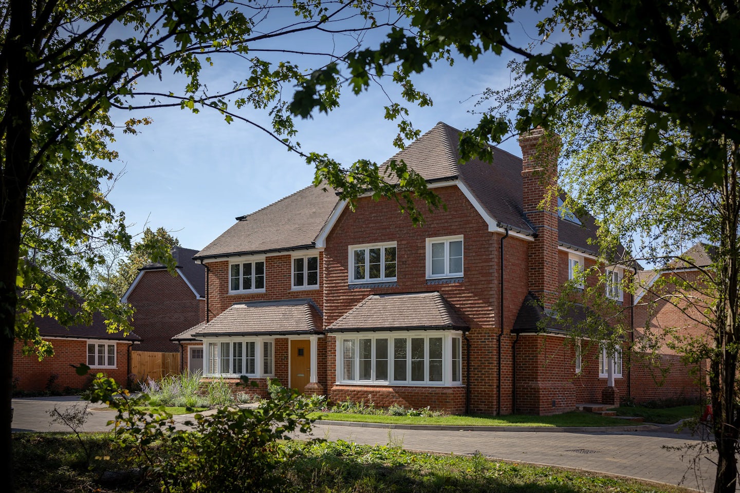 A large, two-story brick house with a pitched roof and multiple windows. Above the main entrance is a circular window, framed by a small porch. The house is surrounded by trees and greenery, suggesting a suburban or semi-rural setting. A paved driveway leads up to the entrance.