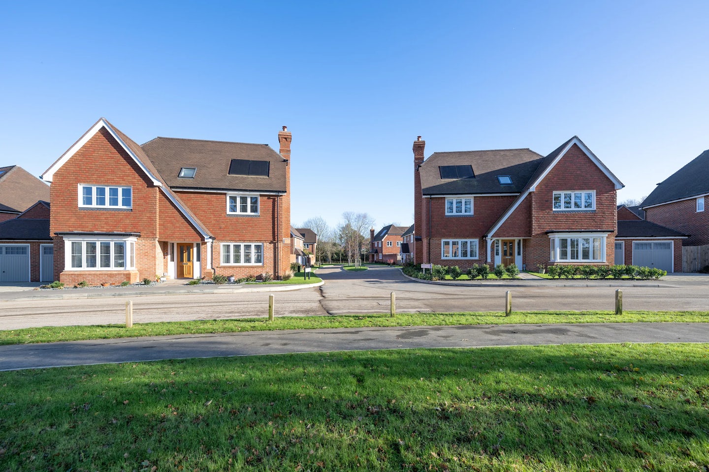 Two large, modern houses with red brick exteriors and pitched roofs. Each house features multiple windows, chimneys, and garages. Positioned on either side of a circular driveway, they are surrounded by a well-maintained lawn. A paved road runs in front of the houses, and the sky is clear and blue, indicating a sunny day.