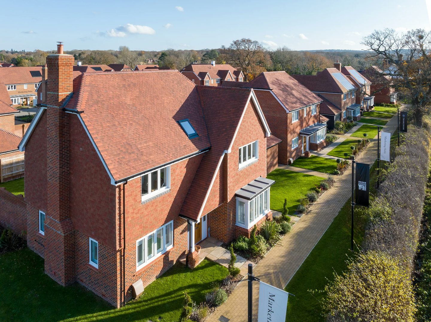 A row of newly built houses in a residential area, featuring red brick exteriors and red tiled roofs. Each house has a well-maintained front lawn and a paved walkway leading to the entrance. The houses are arranged in a neat line along the walkway, which is bordered by a hedge on one side. Signs along the walkway include one that reads "Marketing Suite." The image is taken from an elevated perspective, showcasing modern residential architecture and urban planning.