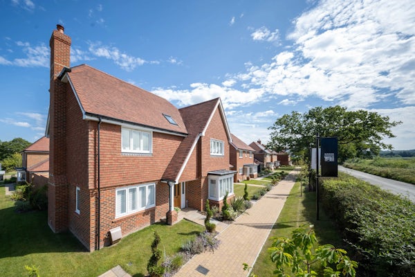 A row of modern detached houses with red brick exteriors and pitched roofs, lined along a paved walkway with landscaped gardens. A signpost is visible near the road, and the sky is partly cloudy. The scene portrays a well-maintained suburban neighbourhood.