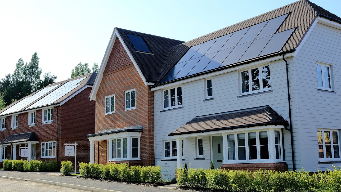 A row of modern houses with solar panels on their roofs. The houses feature a mix of brick and white siding exteriors, with neatly trimmed hedges lining the front. The street is clean and well-maintained, emphasizing the integration of renewable energy in contemporary residential design.