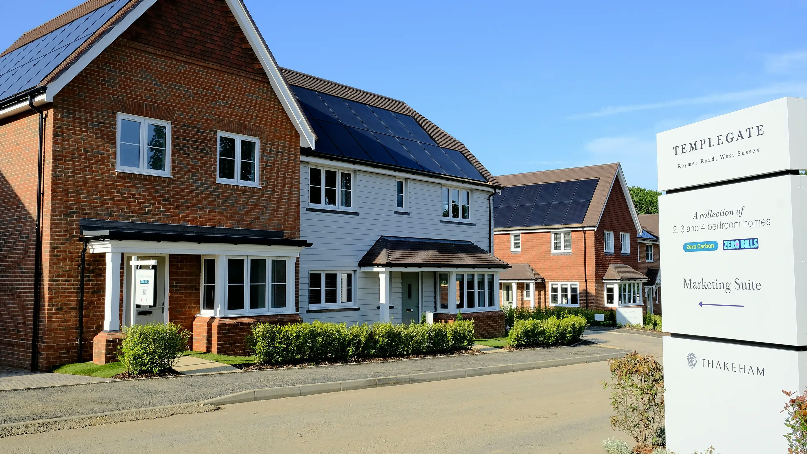 A residential neighbourhood featuring modern homes with brick and white panel exteriors, each equipped with solar panels on the roofs. A prominent signboard in the foreground reads: TEMPLEGATE, Keymer Road, West Sussex A collection of 2, 3, and 4 bedroom homes Zero Carbon ZERO BILLS Marketing Suite ← At the bottom of the signboard, the THAKEHAM logo is displayed. The scene is well-lit, with clear skies and neatly maintained landscaping.
