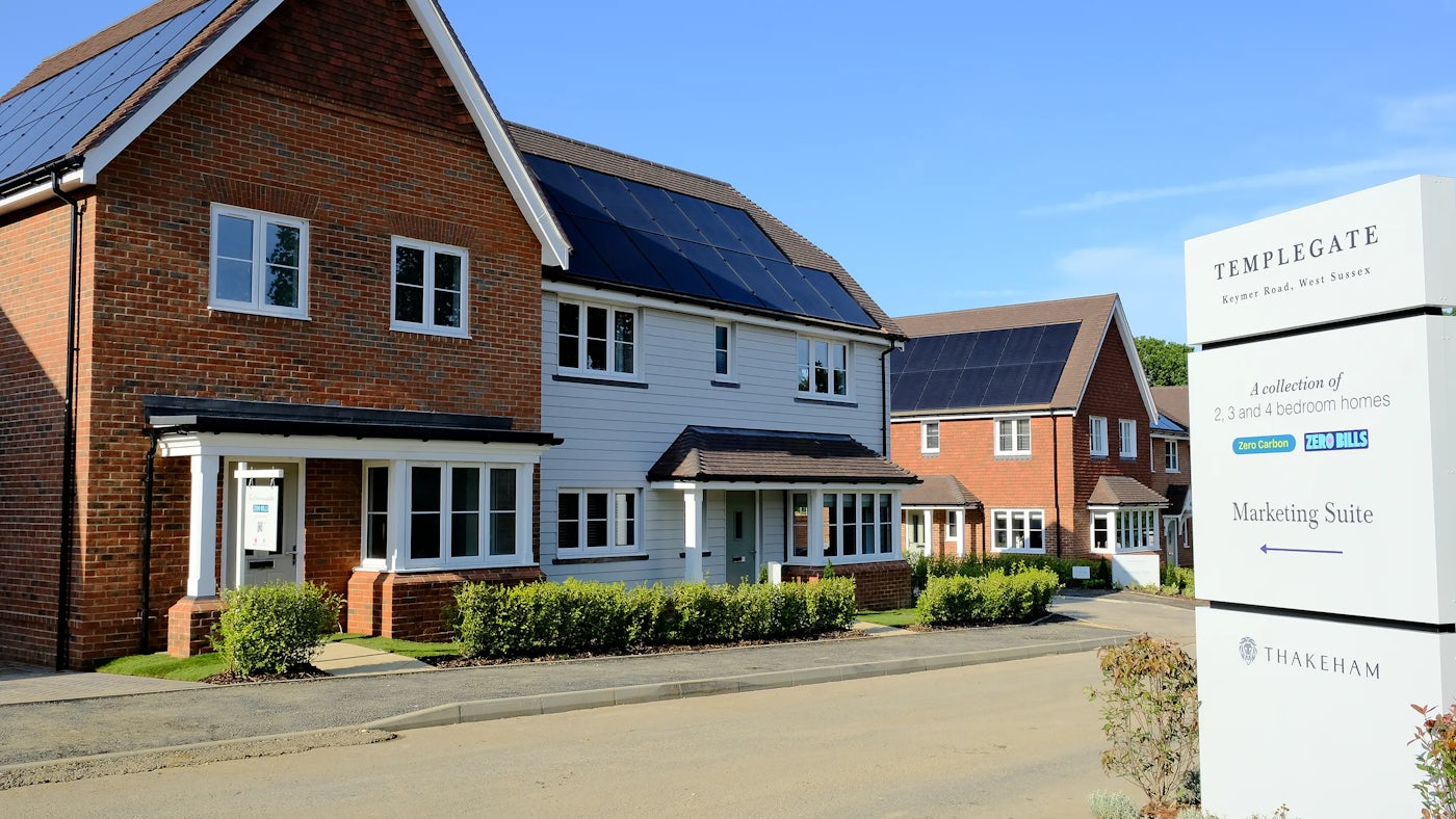 A residential neighbourhood featuring modern homes with brick and white panel exteriors, each equipped with solar panels on the roofs. A prominent signboard in the foreground reads: TEMPLEGATE, Keymer Road, West Sussex A collection of 2, 3, and 4 bedroom homes Zero Carbon ZERO BILLS Marketing Suite ← At the bottom of the signboard, the THAKEHAM logo is displayed. The scene is well-lit, with clear skies and neatly maintained landscaping.