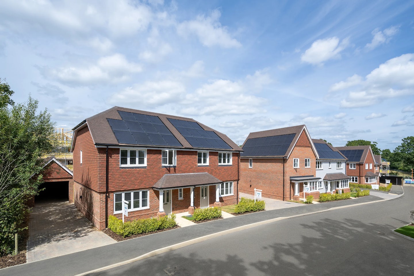 A row of modern, two-story brick houses with solar panels on their roofs lines a well-maintained street. The sky is clear blue with scattered clouds. The presence of solar panels suggests a focus on renewable energy and sustainability.
