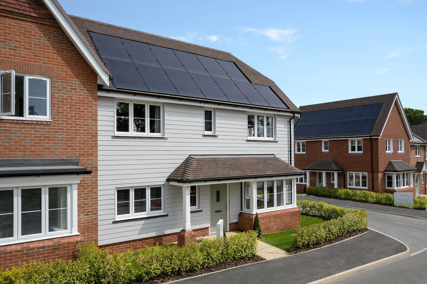 A residential neighbourhood featuring modern two-story houses with brick and white siding exteriors. Solar panels are installed on the rooftops, emphasizing a commitment to renewable energy. The area is well-maintained, with neatly trimmed hedges and a clean, curved road.