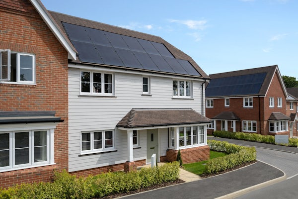 A residential neighbourhood featuring modern two-story houses with brick and white siding exteriors. Solar panels are installed on the rooftops, emphasizing a commitment to renewable energy. The area is well-maintained, with neatly trimmed hedges and a clean, curved road.