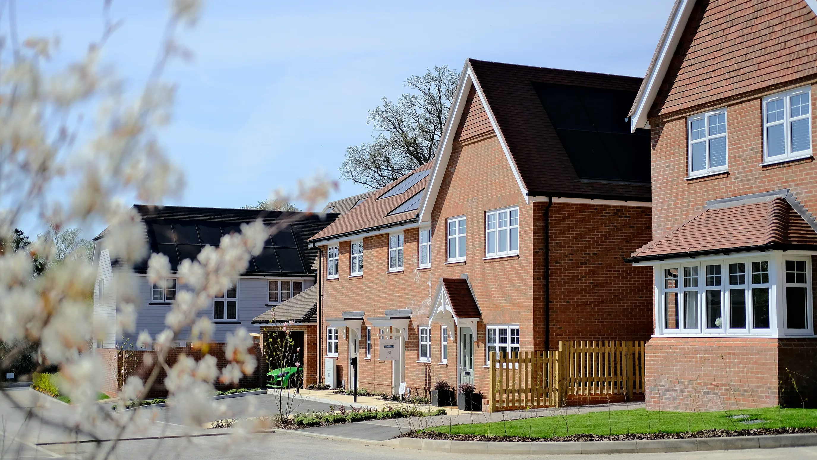 A row of modern, two-story houses with brick exteriors and pitched roofs. Each house has white-framed windows and small front gardens with greenery. A wooden fence separates the properties. In the foreground, blurred branches with white flowers add a natural element to the scene.
