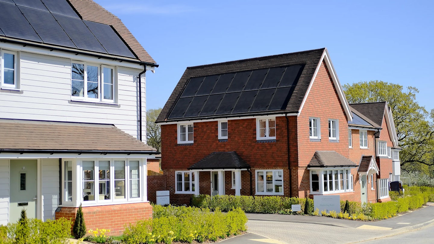 Two modern houses with solar panels on their roofs. The house on the left has a white exterior with a brick base and a large bay window, while the house on the right is red brick with multiple windows, including a bay window. Both houses are surrounded by neatly trimmed hedges and a paved driveway, reflecting a suburban residential setting with a focus on renewable energy and sustainability.