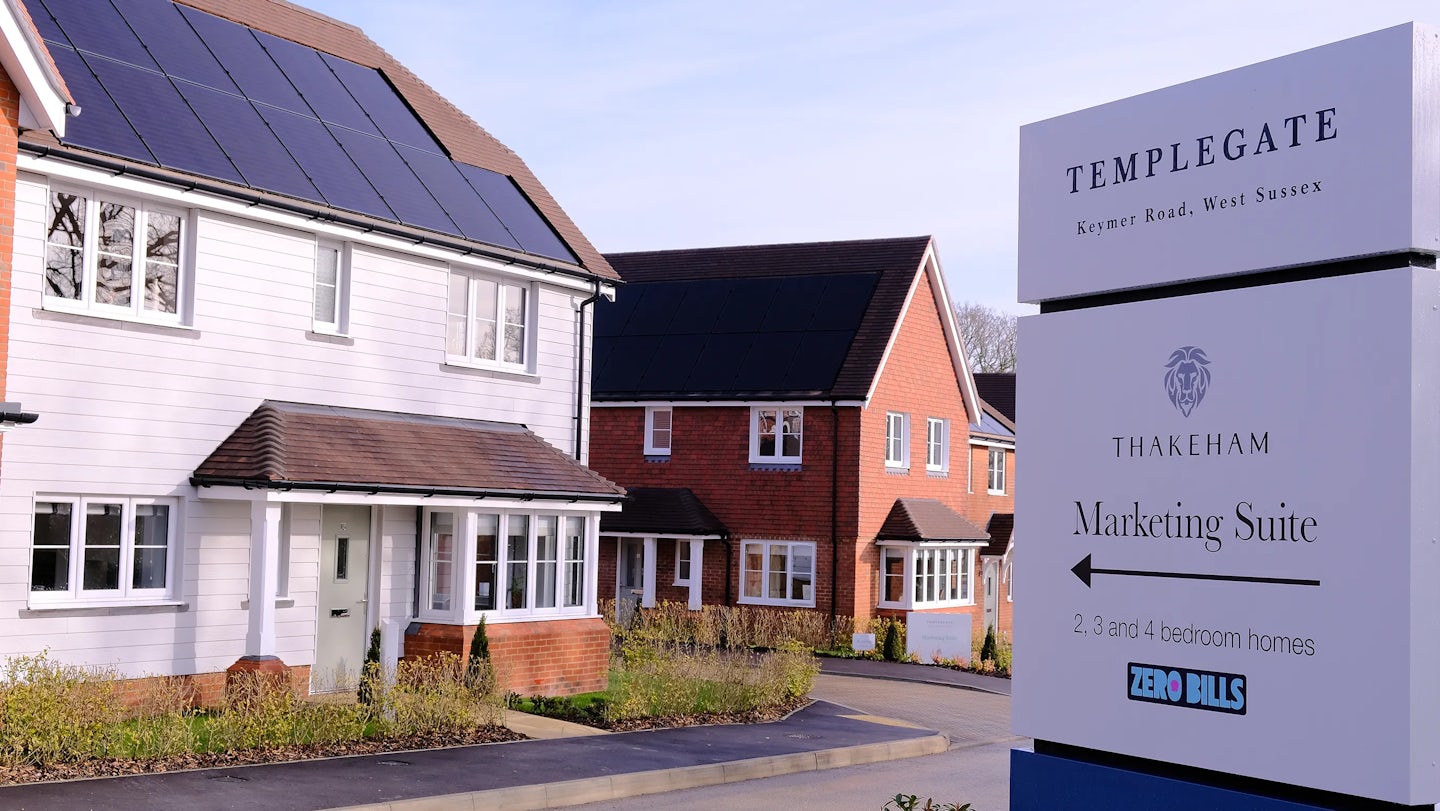 A marketing sign in the foreground reads: TEMPLEGATE Keymer Road, West Sussex THAKEHAM Marketing Suite 2, 3, and 4-bedroom homes ZERO BILLS Behind the sign, a row of modern houses is visible, some featuring solar panels on their roofs. The setting indicates a new residential development focused on energy efficiency.