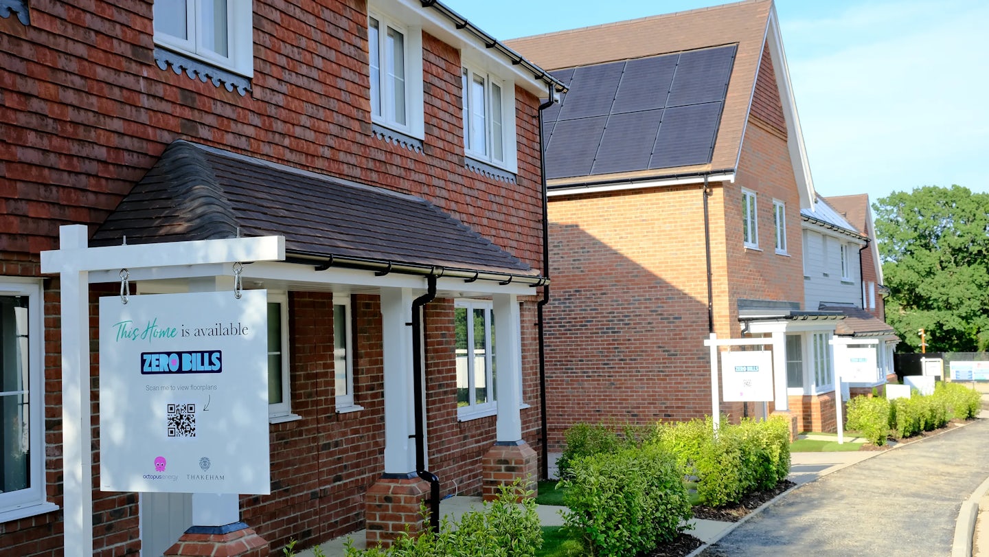 A row of modern brick houses with "ZERO BILLS" signs in front, indicating energy-efficient homes. One house features solar panels on the roof. The signs display QR codes and logos for "Octopus Energy" and "Thakeham," suggesting involvement in sustainable housing development.