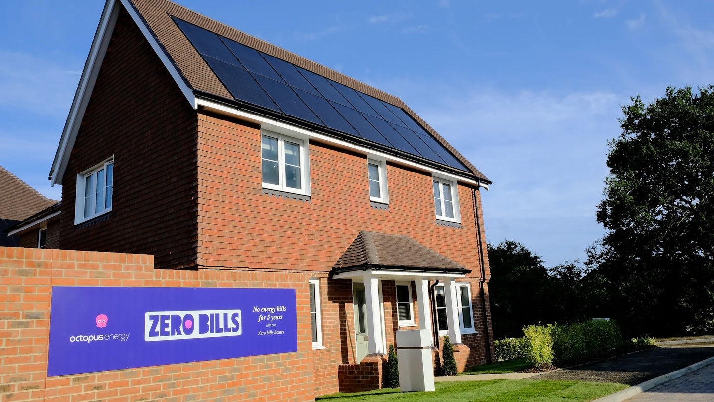 A modern brick house with solar panels on its roof. The house has white-framed windows and a small porch. A sign on the brick wall next to the house reads: "octopus energy ZERO BILLS No energy bills for 5 years Zero bills homes."