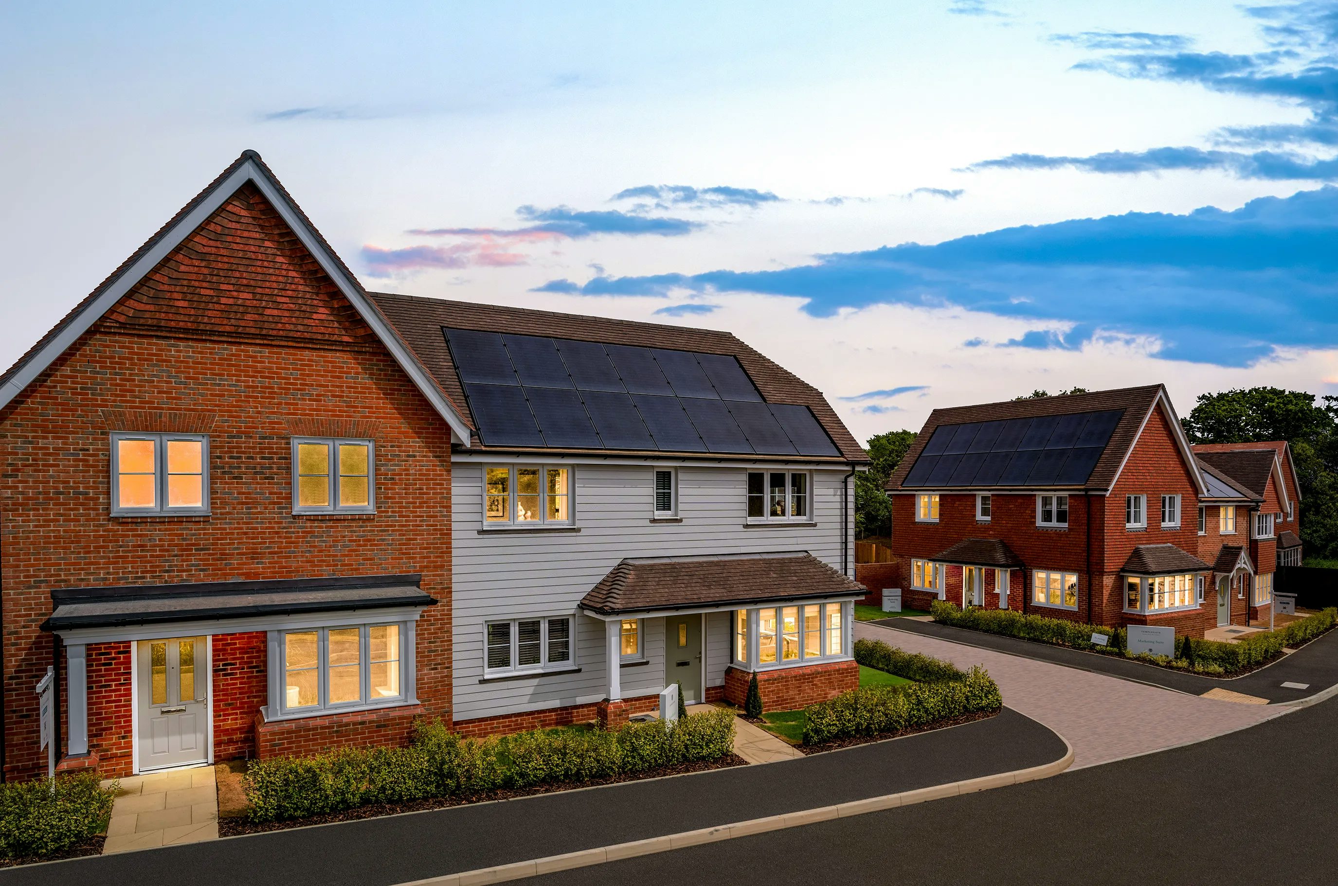 A residential neighbourhood at dusk, featuring modern houses with a mix of brick and siding exteriors. Some rooftops are equipped with solar panels, highlighting sustainable living. The sky displays a blend of blue and pink hues, casting a warm light over the well-maintained landscaping and paved roads.