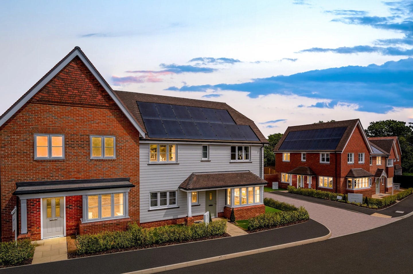 A residential neighbourhood at dusk, featuring modern houses with a mix of brick and siding exteriors. Some rooftops are equipped with solar panels, highlighting sustainable living. The sky displays a blend of blue and pink hues, casting a warm light over the well-maintained landscaping and paved roads.
