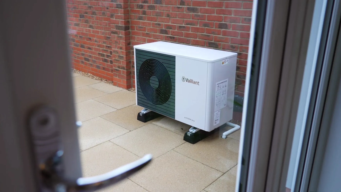 A Vaillant outdoor heat pump unit is mounted on two black supports on a paved surface next to a brick wall. The white casing features a large circular fan on the front, with a label containing text and diagrams on the side. The image is viewed through a glass door from inside a building.